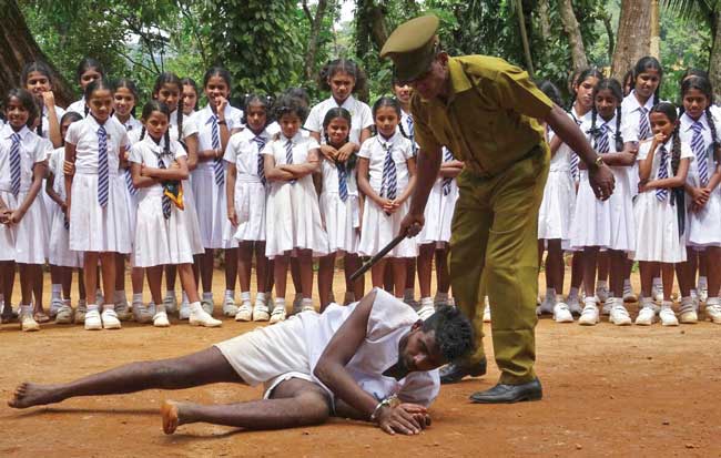 An actor dressed as a guard pretends to beat an actor dressed as a prisoner in an outdoor space while an audience of uniformed schoolgirls watch.
