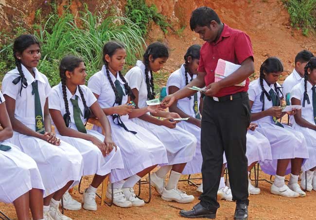 A missionary hands out gospel literature to a row of uniformed schoolgirls seated outside.